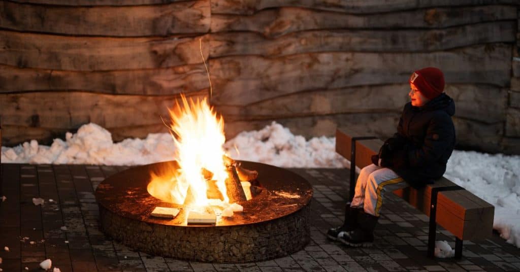 Person sits on a wooden bench beside a circular fire pit in snow.