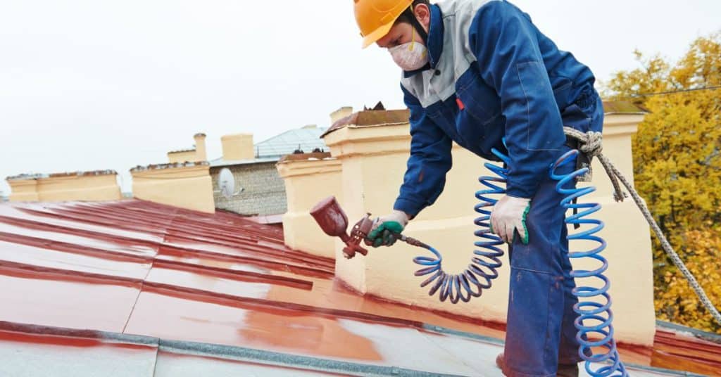 Worker painting metal roof surface using spray gun and coiled hose
