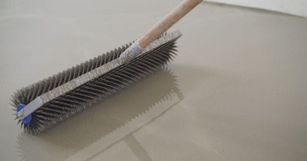 Stiff-bristle roller spreads wet self-leveling cement on floor.
