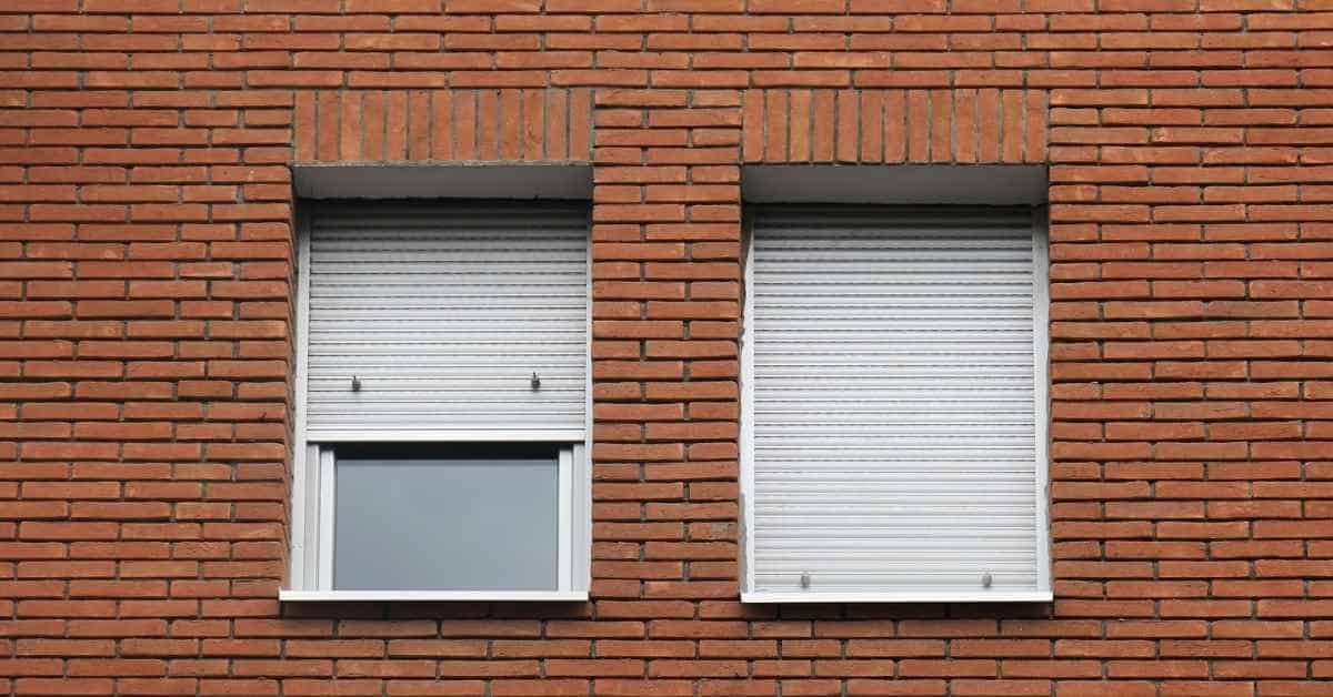 Two basement windows with closed white roller shutters in brick wall