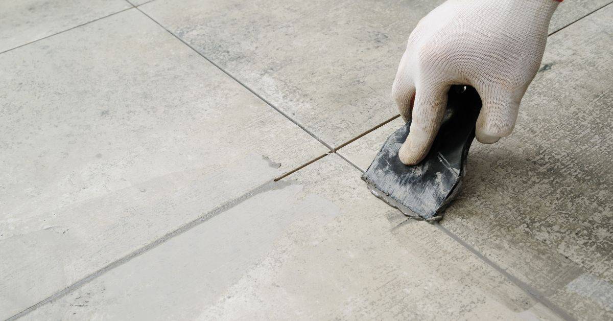 Hand wearing white glove applying grout to tile gap with scraper