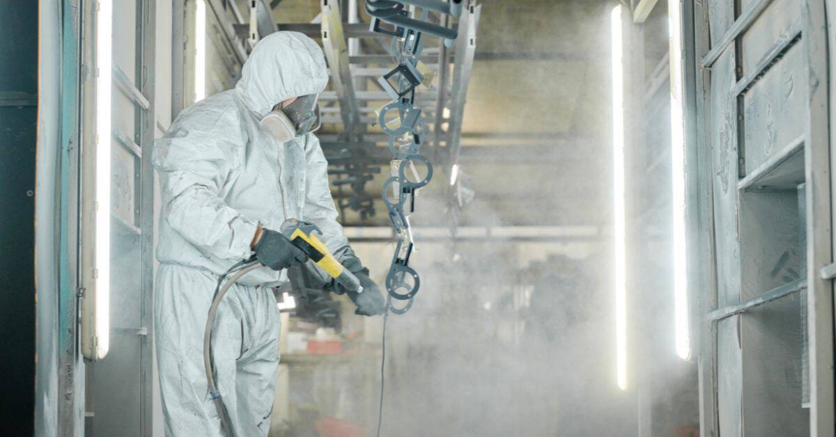 Worker in protective suit spraying coating on metal parts