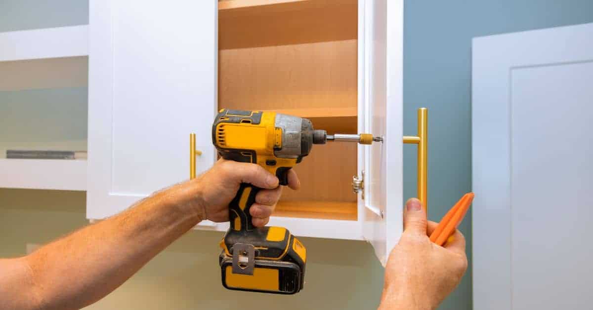 Hands removing gold handle from white cabinet door with drill