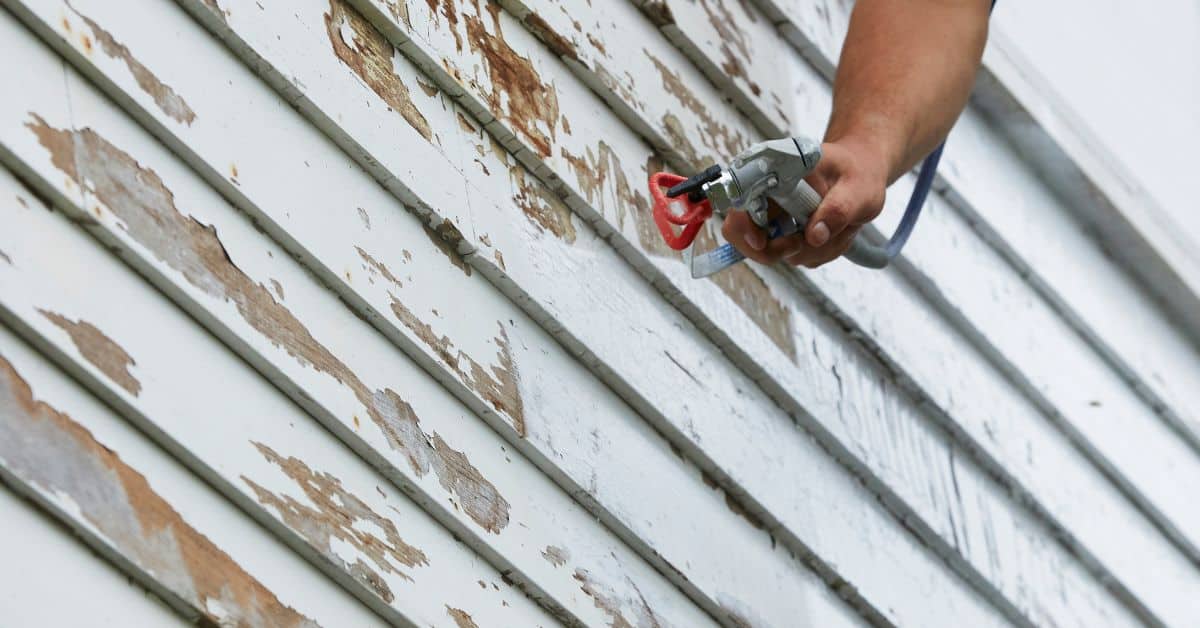 hand holding spray paint gun near peeling white wood siding