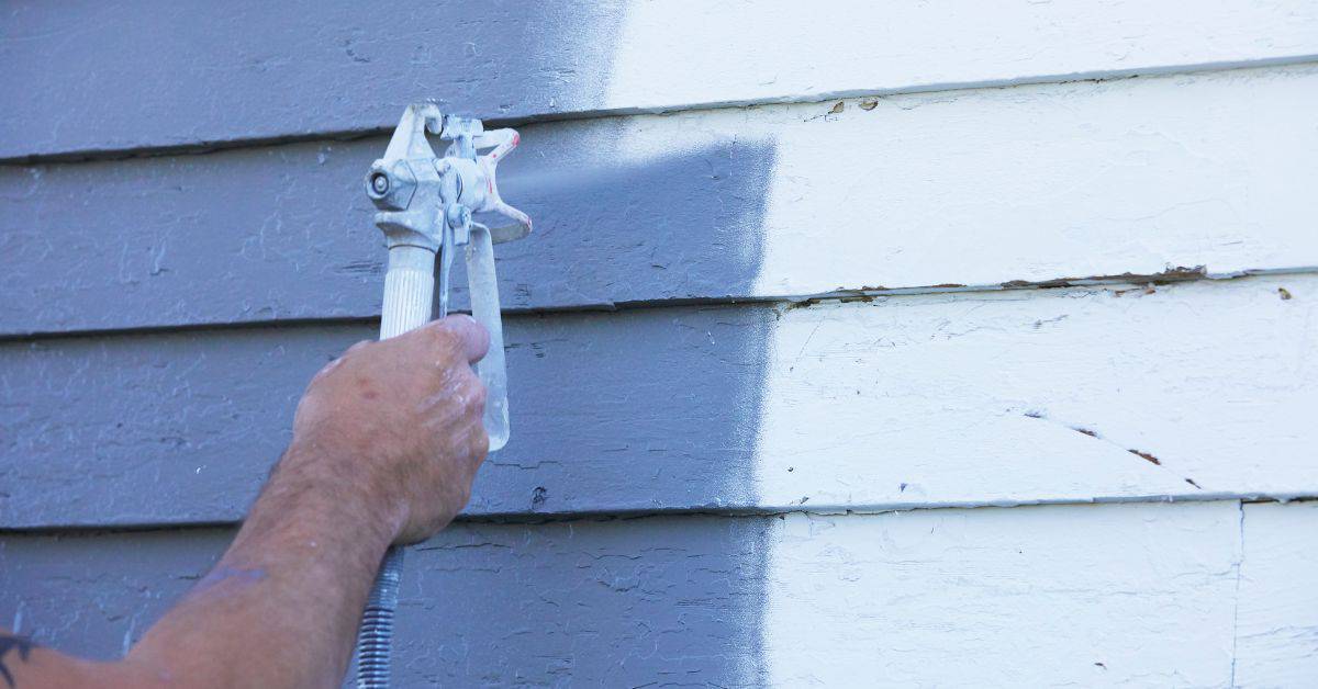 Hand spraying gray paint on white wooden siding