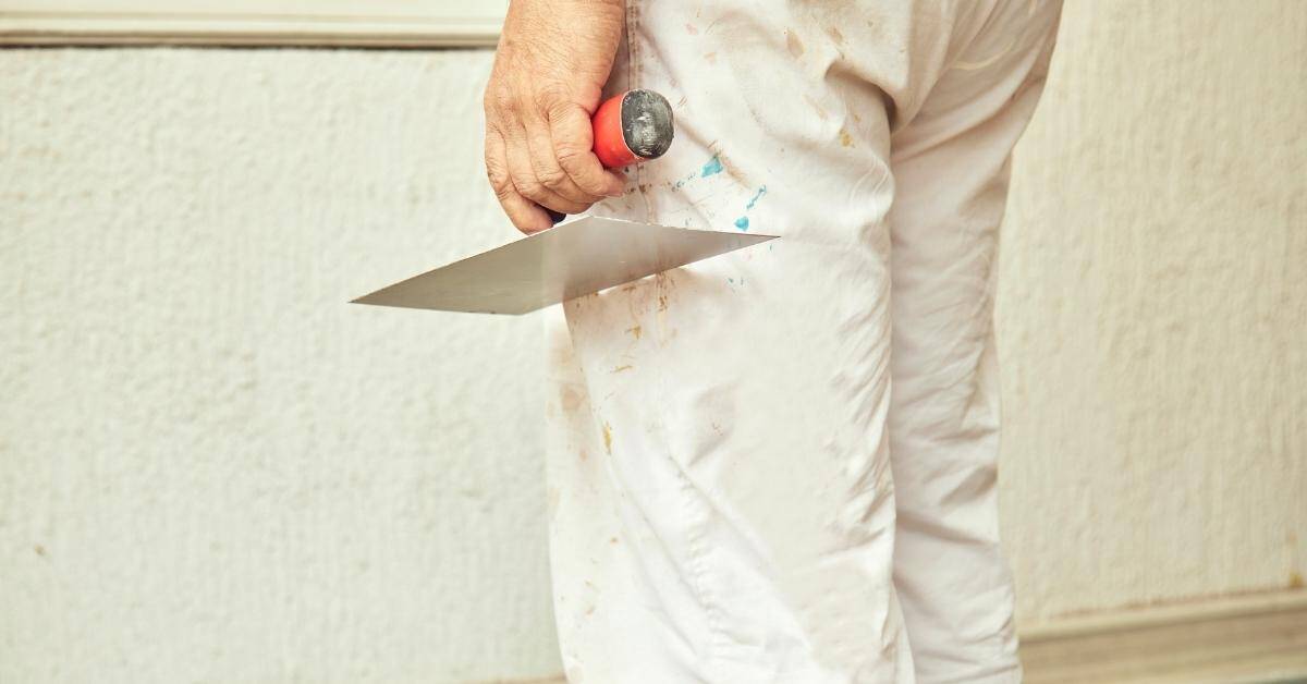 Hand holding plastering trowel with red handle near wall corner