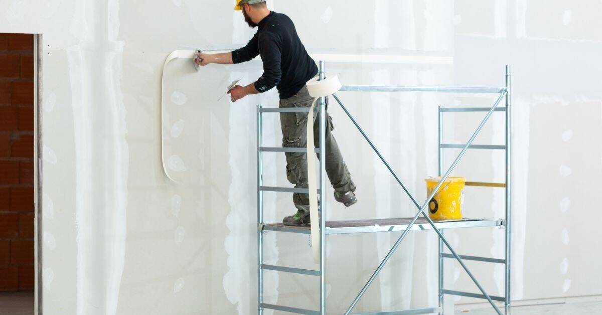Worker applying drywall joint tape on wall while standing on scaffold