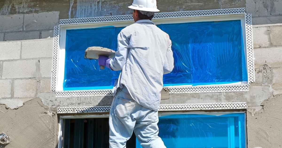 Worker applying stucco near window covered in blue plastic