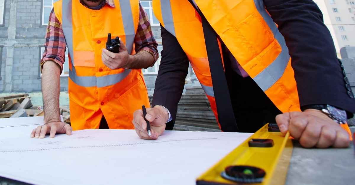 Two construction workers in orange safety vests reviewing blueprints