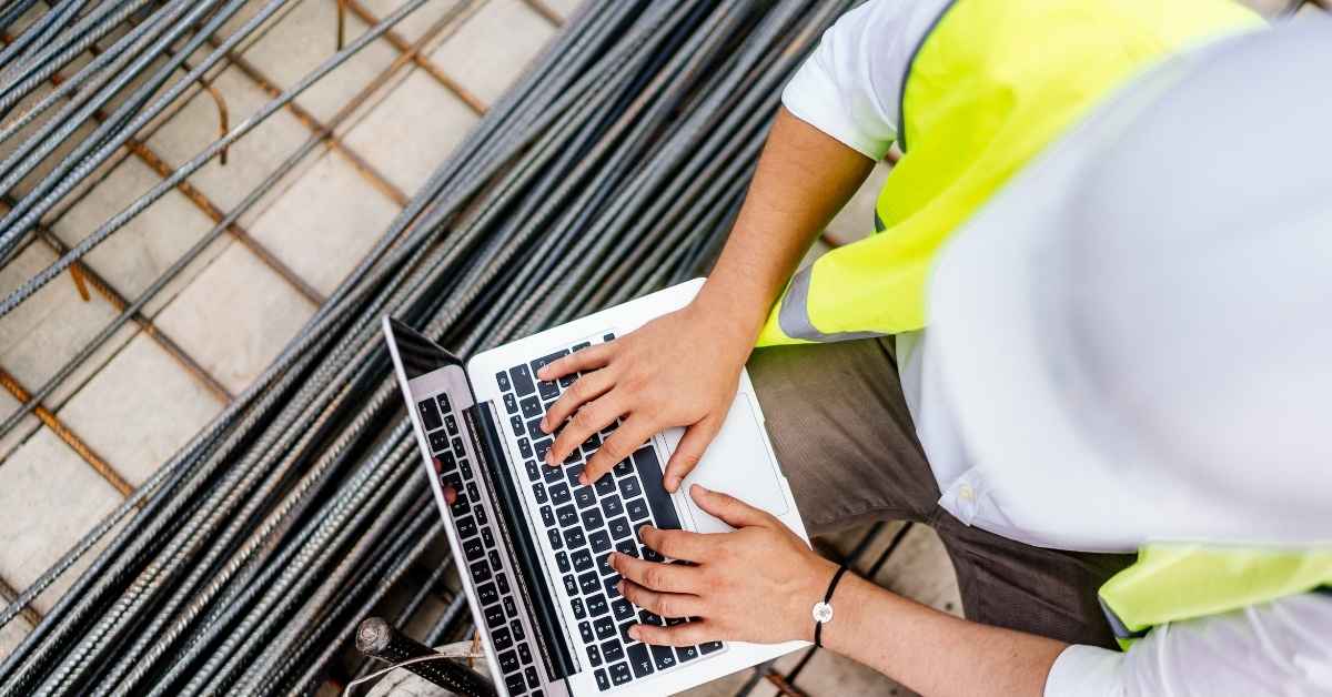 Construction worker typing on laptop beside metal rods