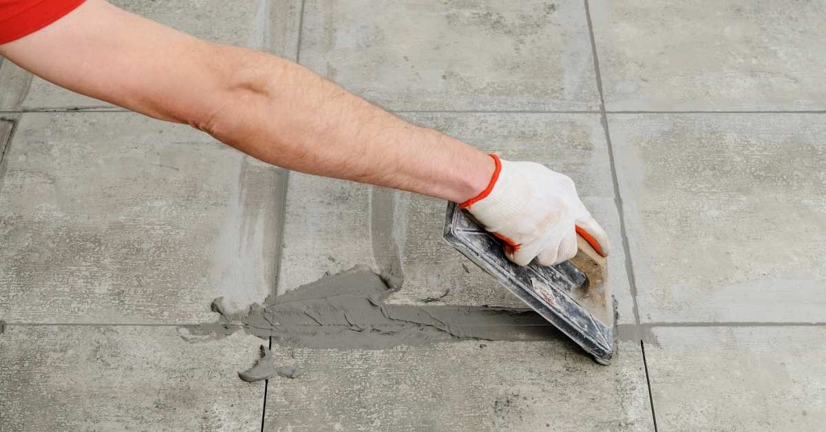 Hand with glove spreading grout over floor tiles with trowel