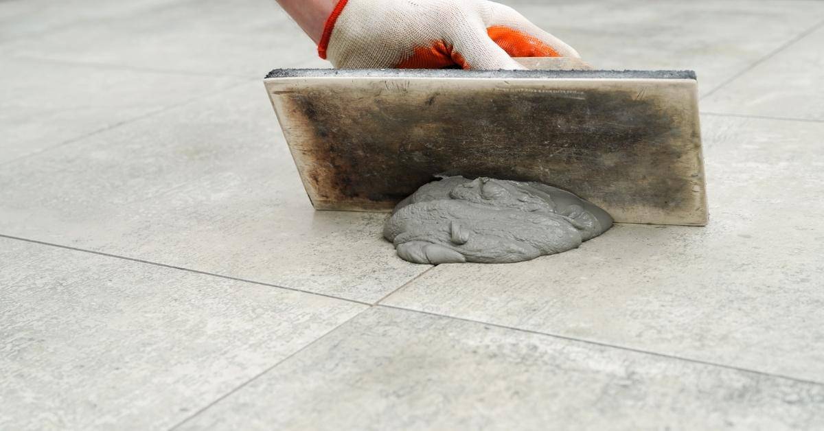 Hand spreading gray grout on tiled floor using float