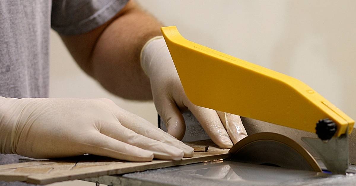 Hands wearing gloves cutting tile with wet saw blade