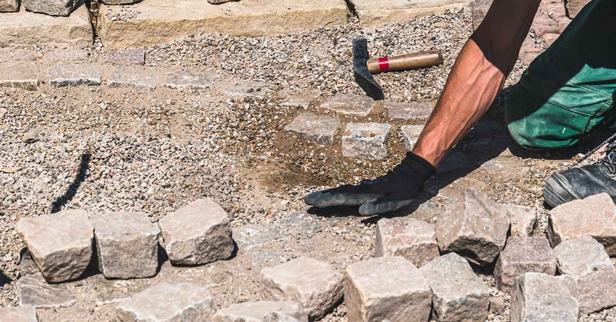 Person's gloved hand arranging cobblestones on gravel base outdoors