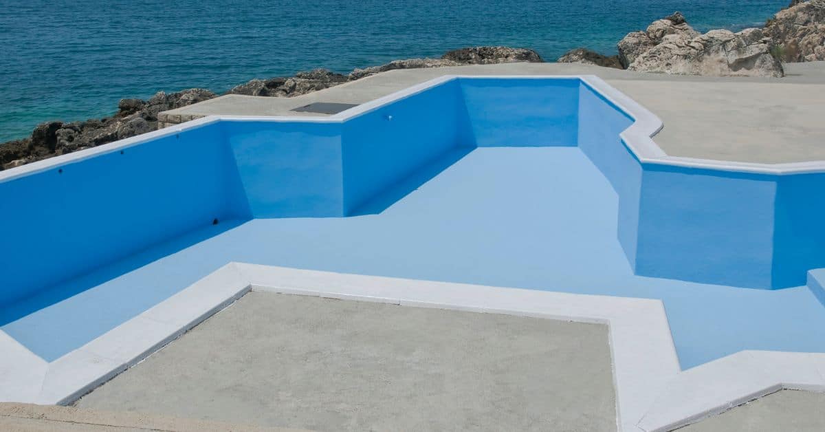 Empty blue painted concrete pool shell near rocky coastline