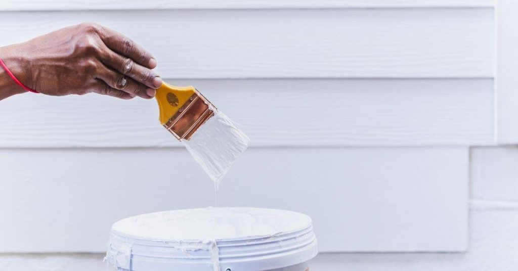 Hand dipping paintbrush into white paint bucket