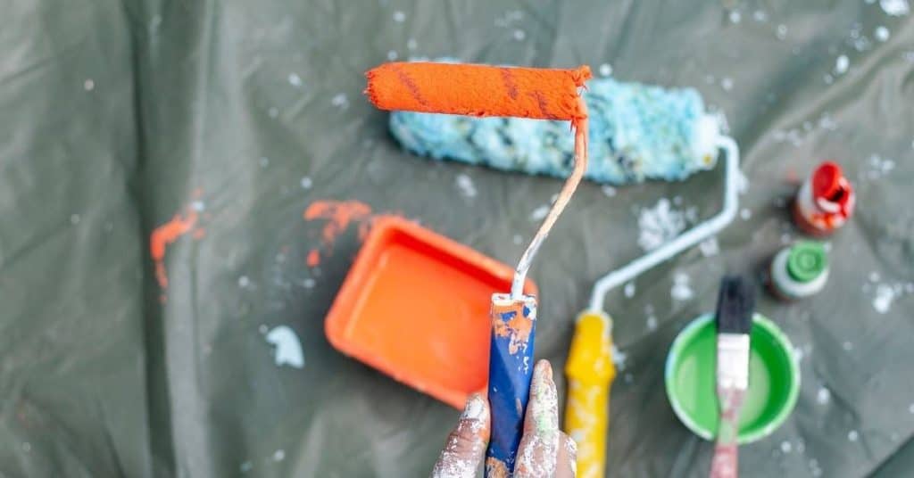 Hand holding paint roller with orange paint over plastic drop cloth