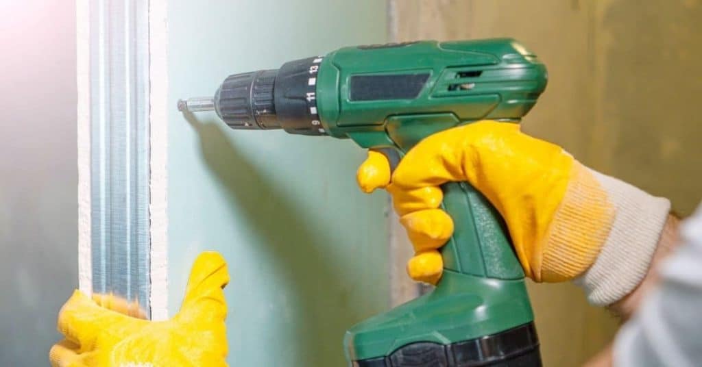 Hand with yellow gloves holding green cordless drill against drywall