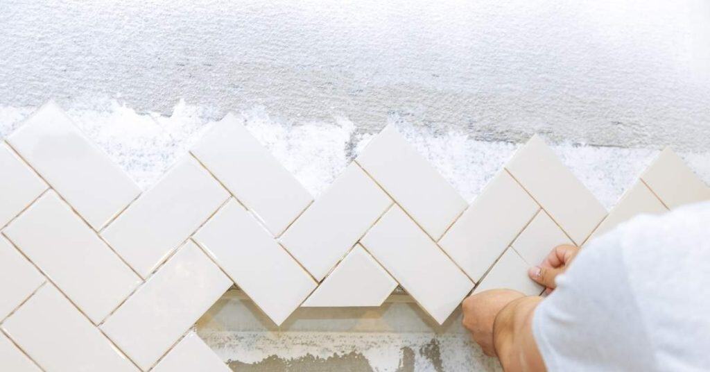 Hands installing white ceramic tiles in herringbone pattern