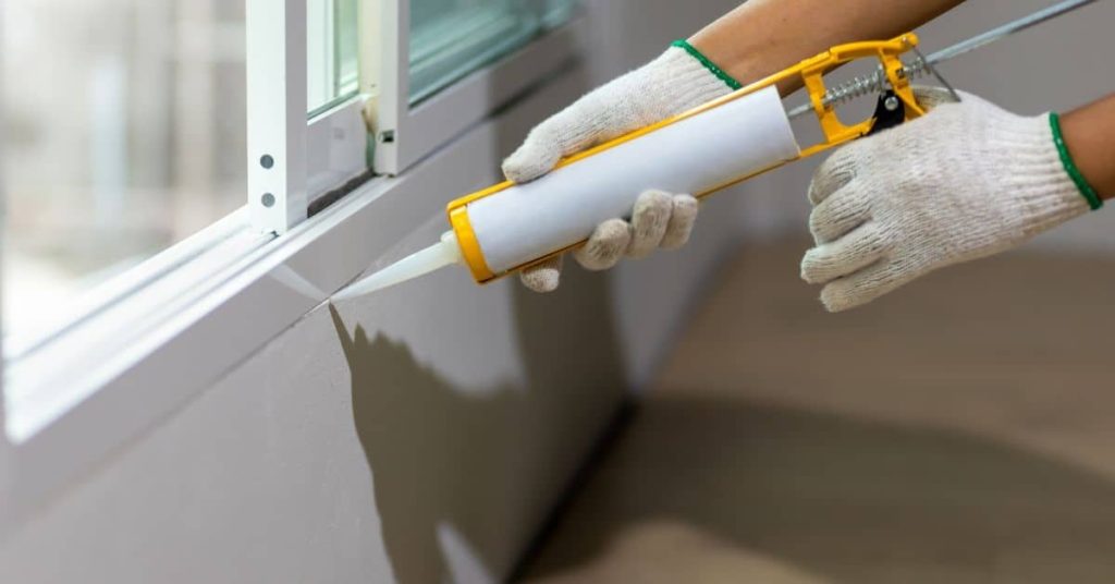 Hands wearing gloves applying white caulk to window frame joint