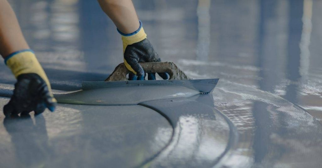 Hands wearing gloves smoothing gray epoxy on floor with trowel