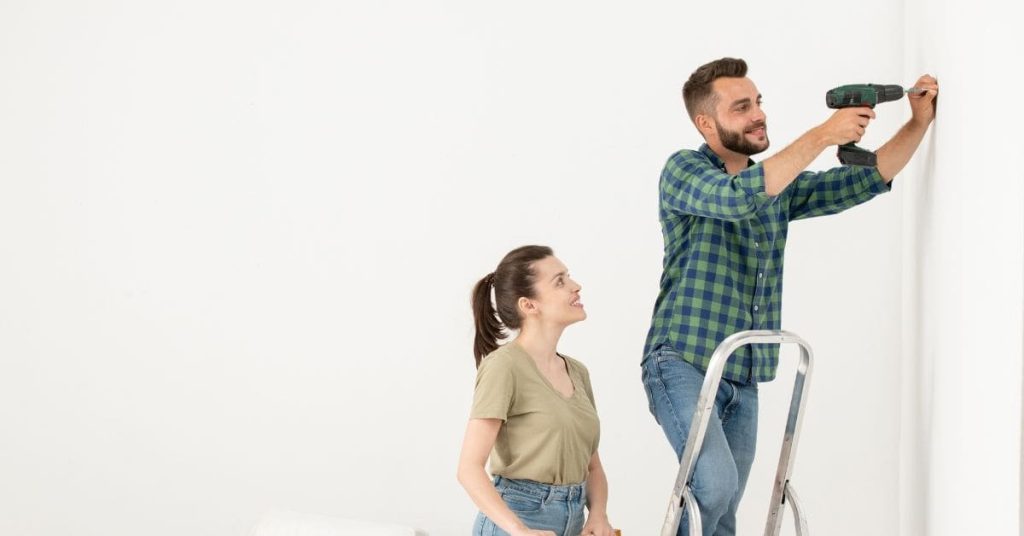 Man using cordless drill on white wall with woman watching