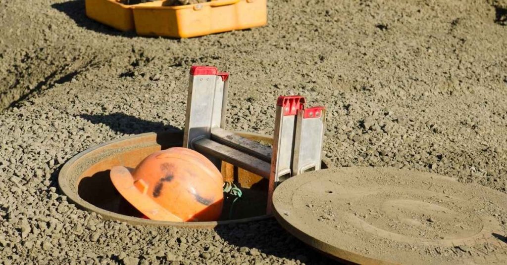 Orange hard hat inside open manhole surrounded by gravel