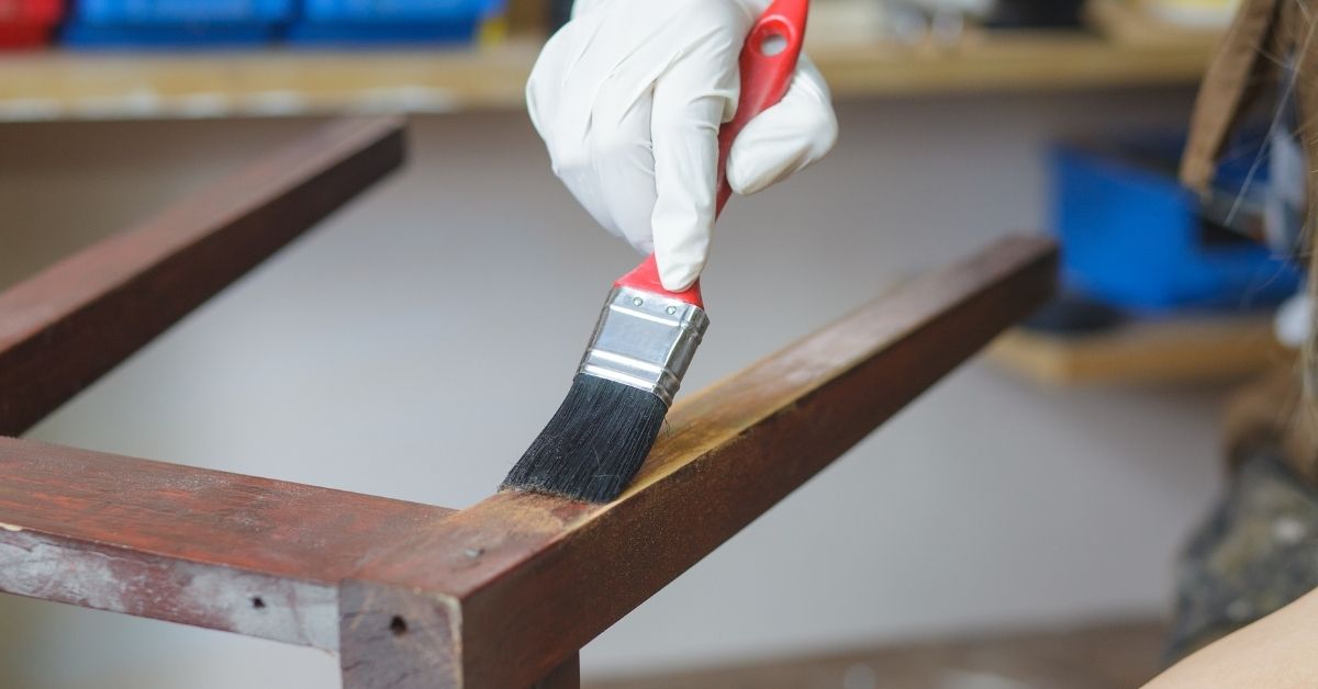 Hand in glove applying varnish with paintbrush on wooden frame