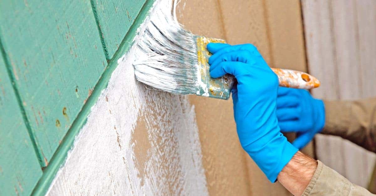 Person wearing blue gloves painting white primer on wood siding