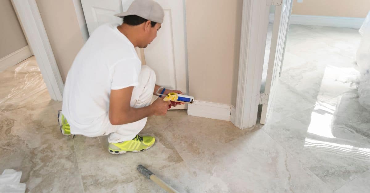 Man applying caulk to baseboards on tile floor