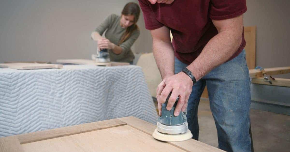 Two people sanding wooden surfaces with electric sanders indoors