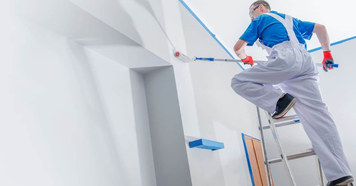 person painting wall white using roller on ladder