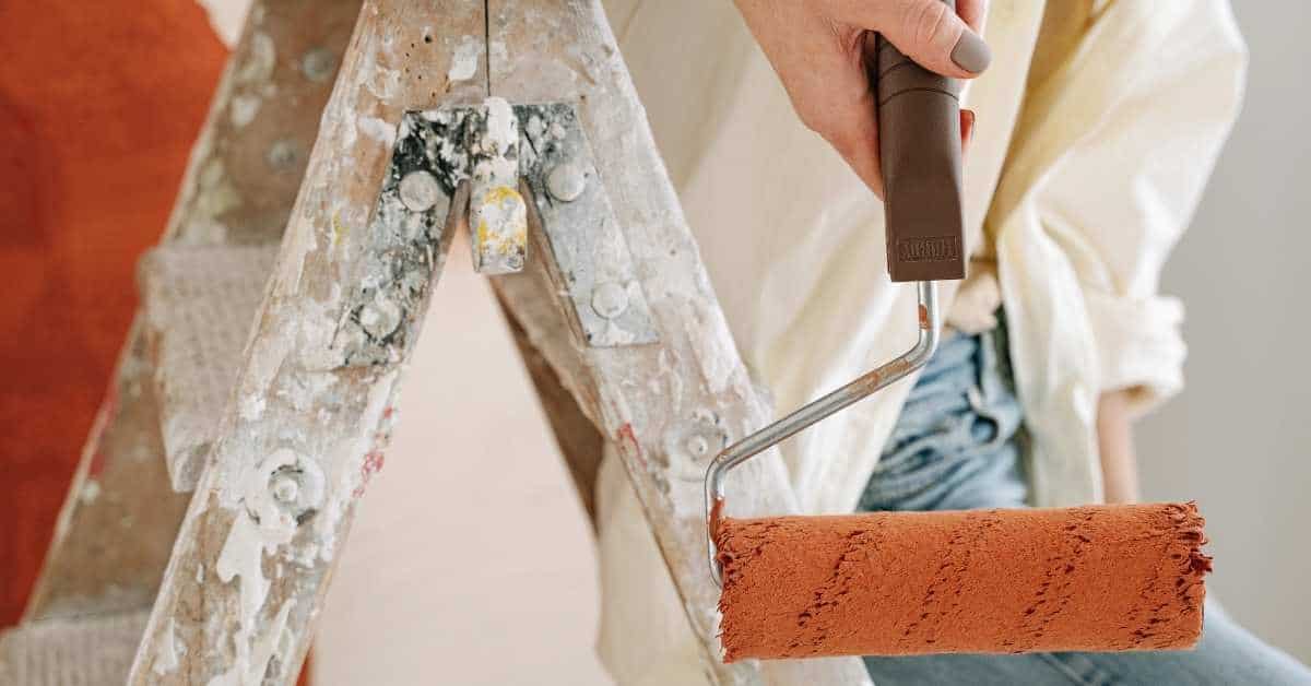 Person holding paint roller with rust-colored paint near paint-splattered ladder