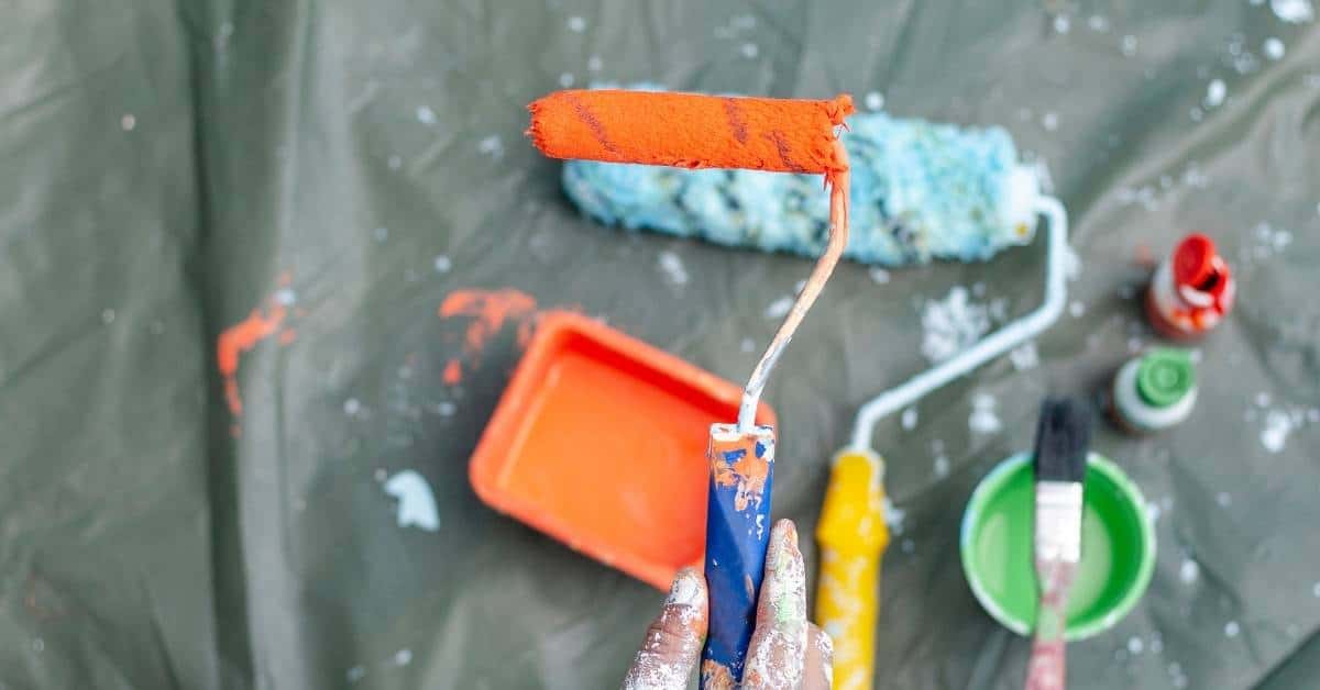 Hand holding paint roller with bright orange paint and supplies