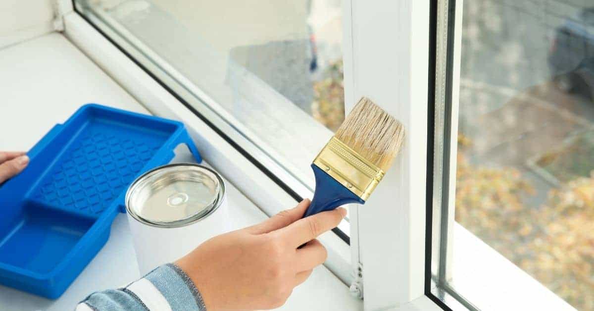 Hand holding paintbrush near white window frame with paint tray