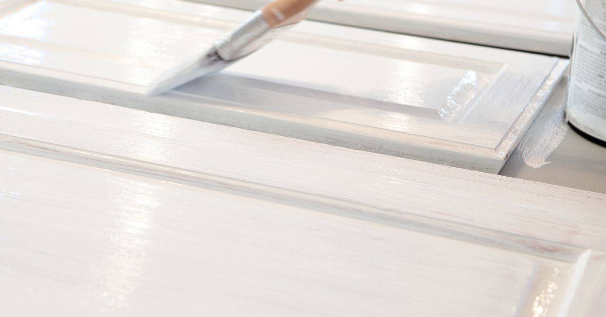 Paintbrush applying white paint on wooden cabinet panels
