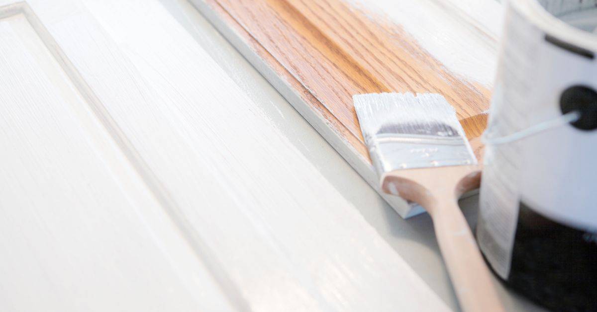 Paintbrush applying white primer to wooden cabinet panel