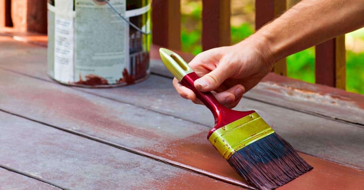 Hand holding paintbrush applying red paint on wooden deck boards