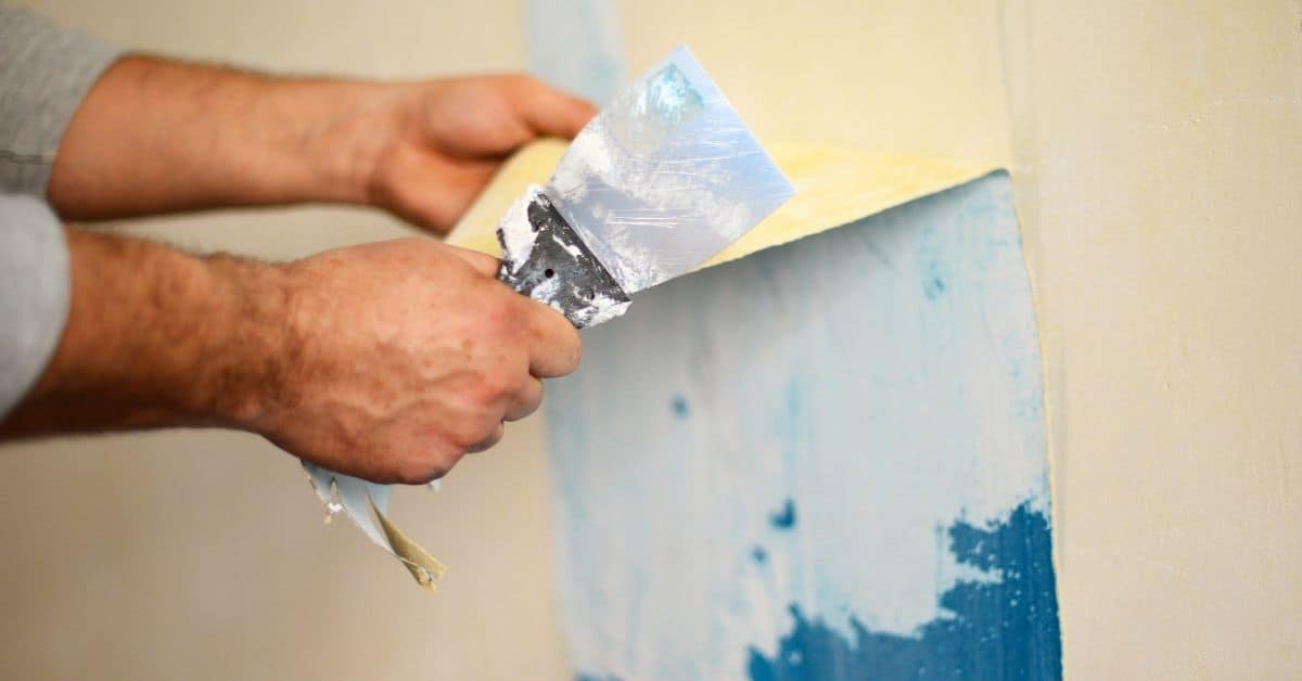 hands scraping blue paint off a wall with a putty knife
