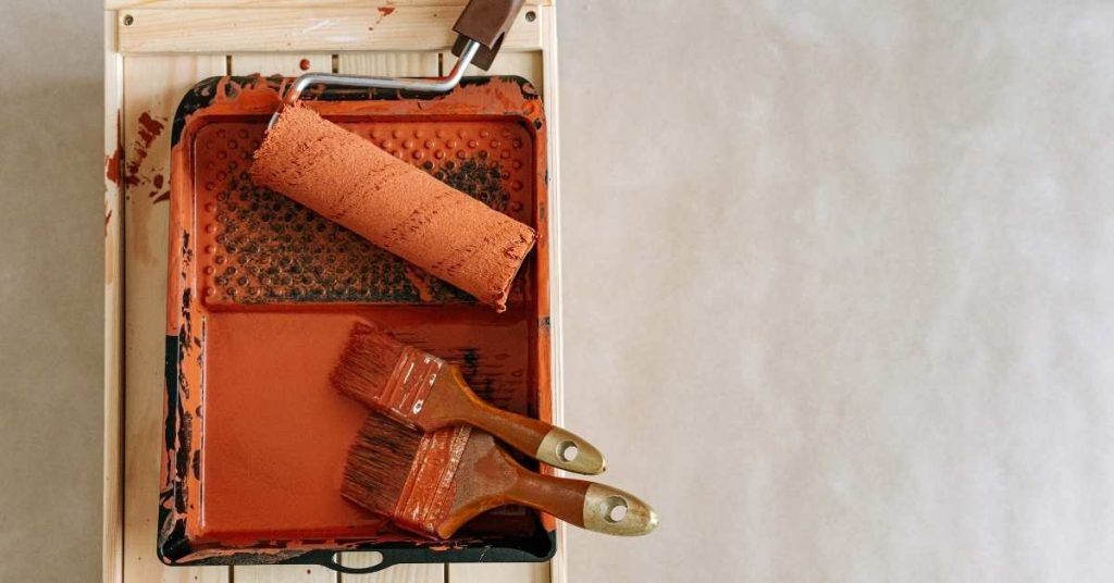 rust-colored paint roller and paintbrushes in tray on wooden surface