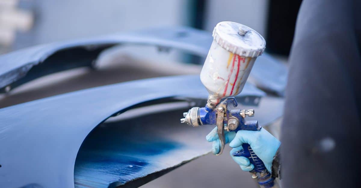 Hand wearing blue glove holding spray paint gun near car surface