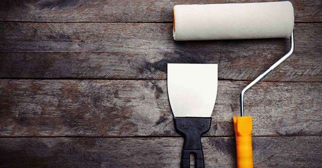 Paint roller with orange handle and metal putty knife on wooden surface