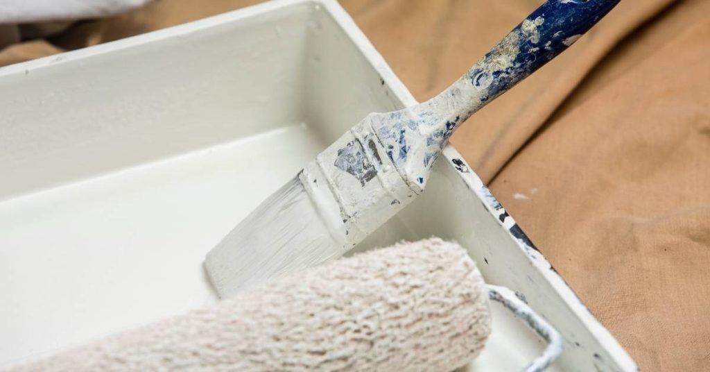 Paintbrush and roller in white paint tray on brown surface