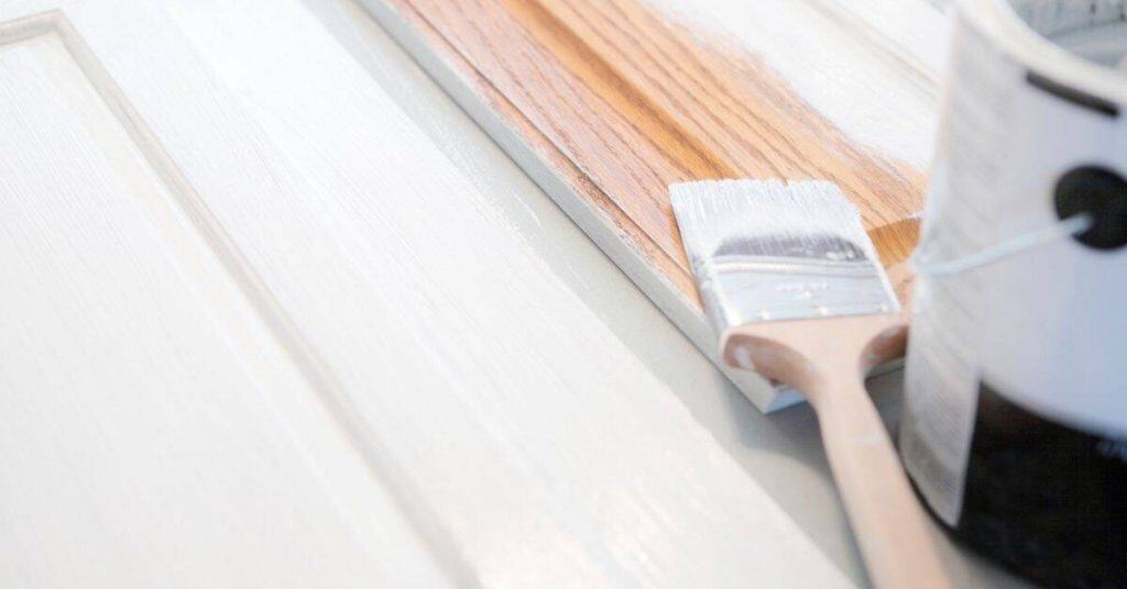 Paintbrush applying white paint to wooden trim