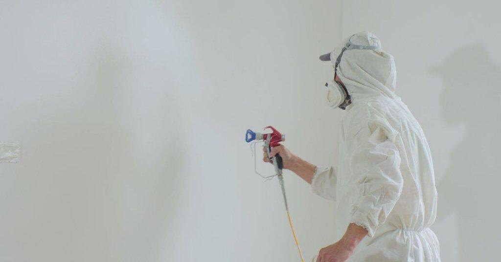 Painter in protective suit spraying white paint on wall