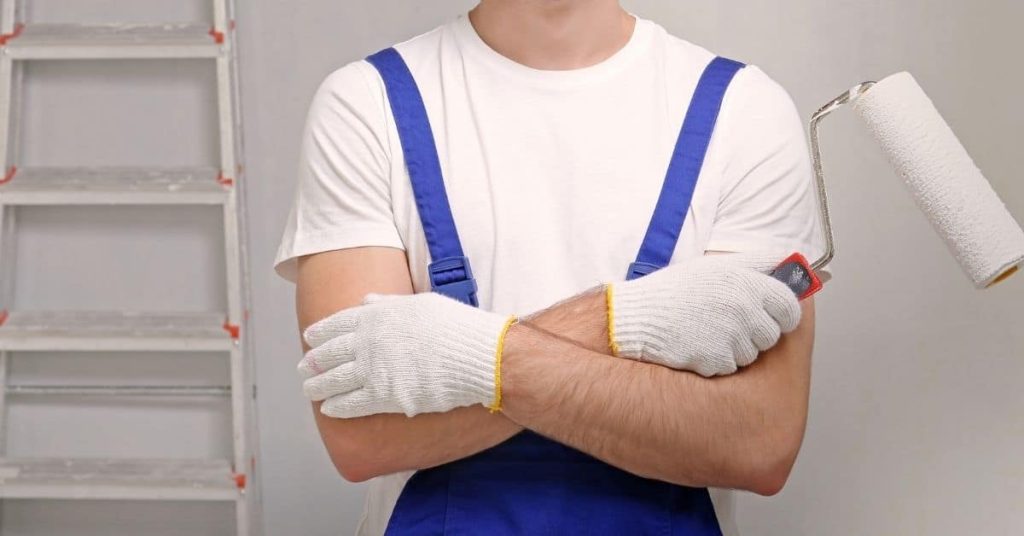 Person holding paint roller wearing gloves and blue overalls