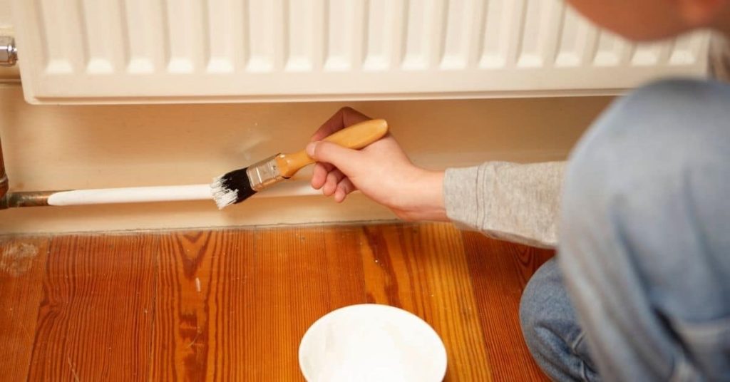 Person painting a metal pipe white near wooden floor and radiator
