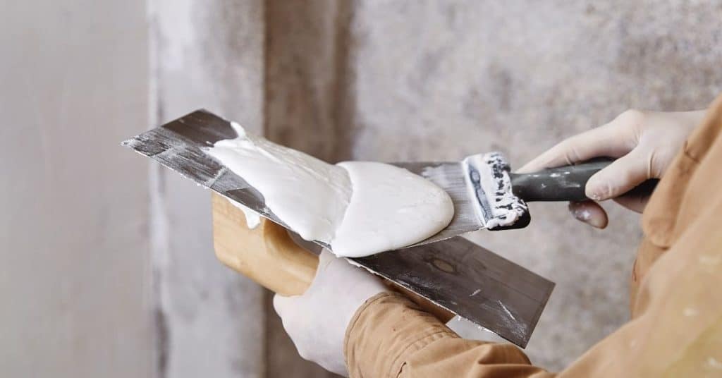 Person spreading white plaster on metal trowel with spatula
