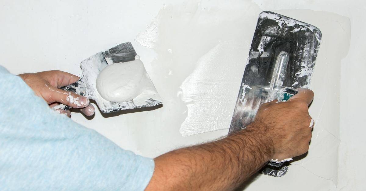 Person applying white plaster on wall using trowel and putty knife