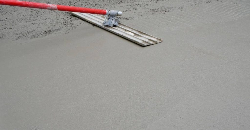 Red-handled concrete screed tool smoothing wet concrete surface
